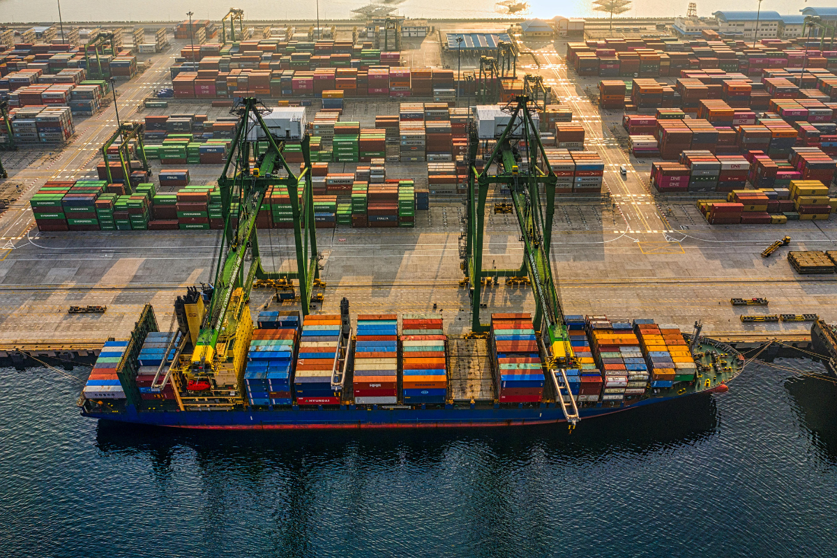 Aerial view of a commercial shipping port with container ship and cargo containers, representing offshore electronics manufacturing supply chain complexity and logistics costs